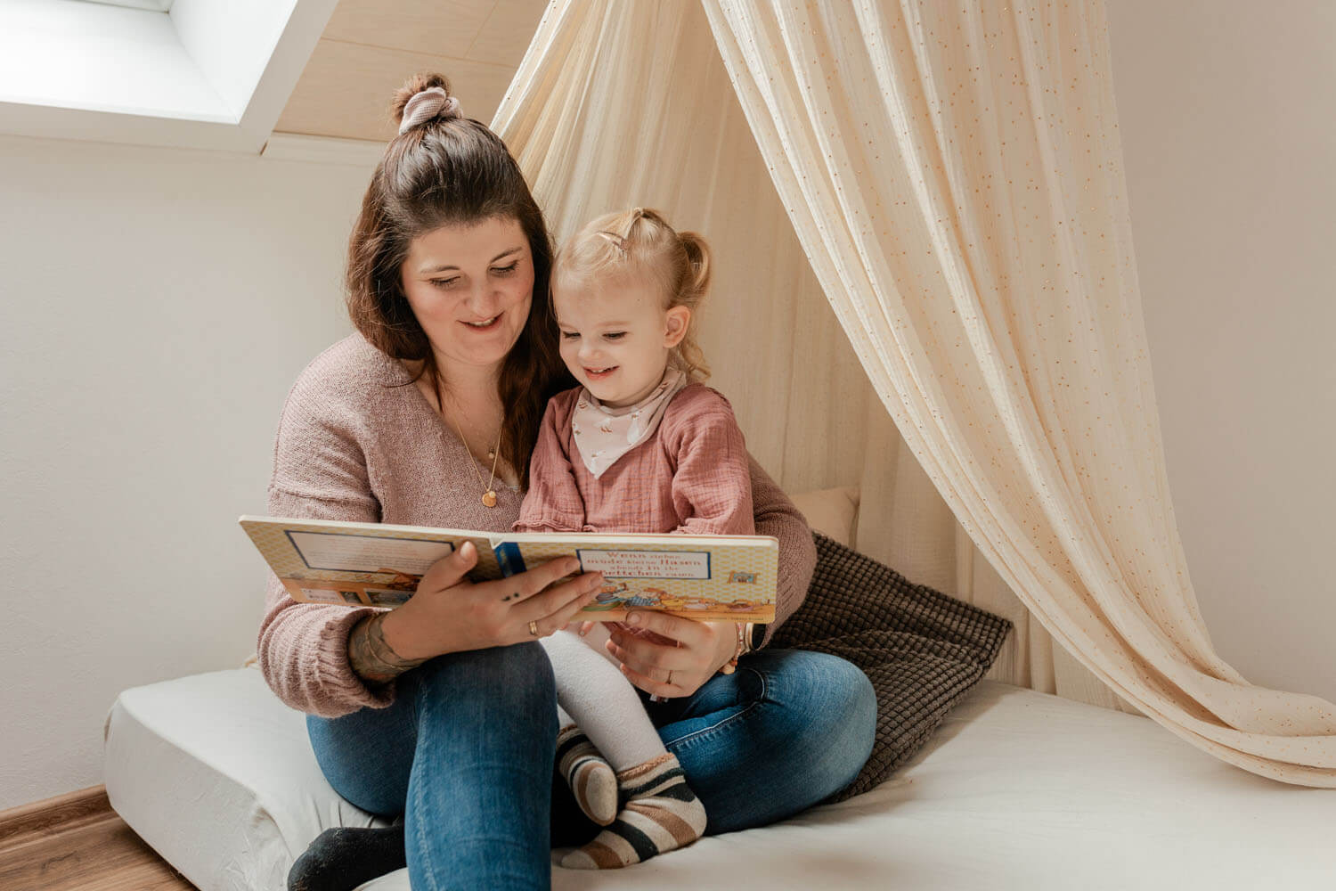 Mama sitzt in der Kuschelecke im Kinderzimmer, ihre Tochter hat sich an sie gekuschelt und liest ihr etwas aus ihrem Lieblingsbuch vor.