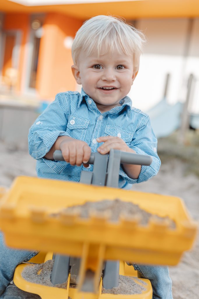 Ein kleiner Junge sitzt im Sandkasten auf einem Spielzeugbagger und buddelt im Sand.